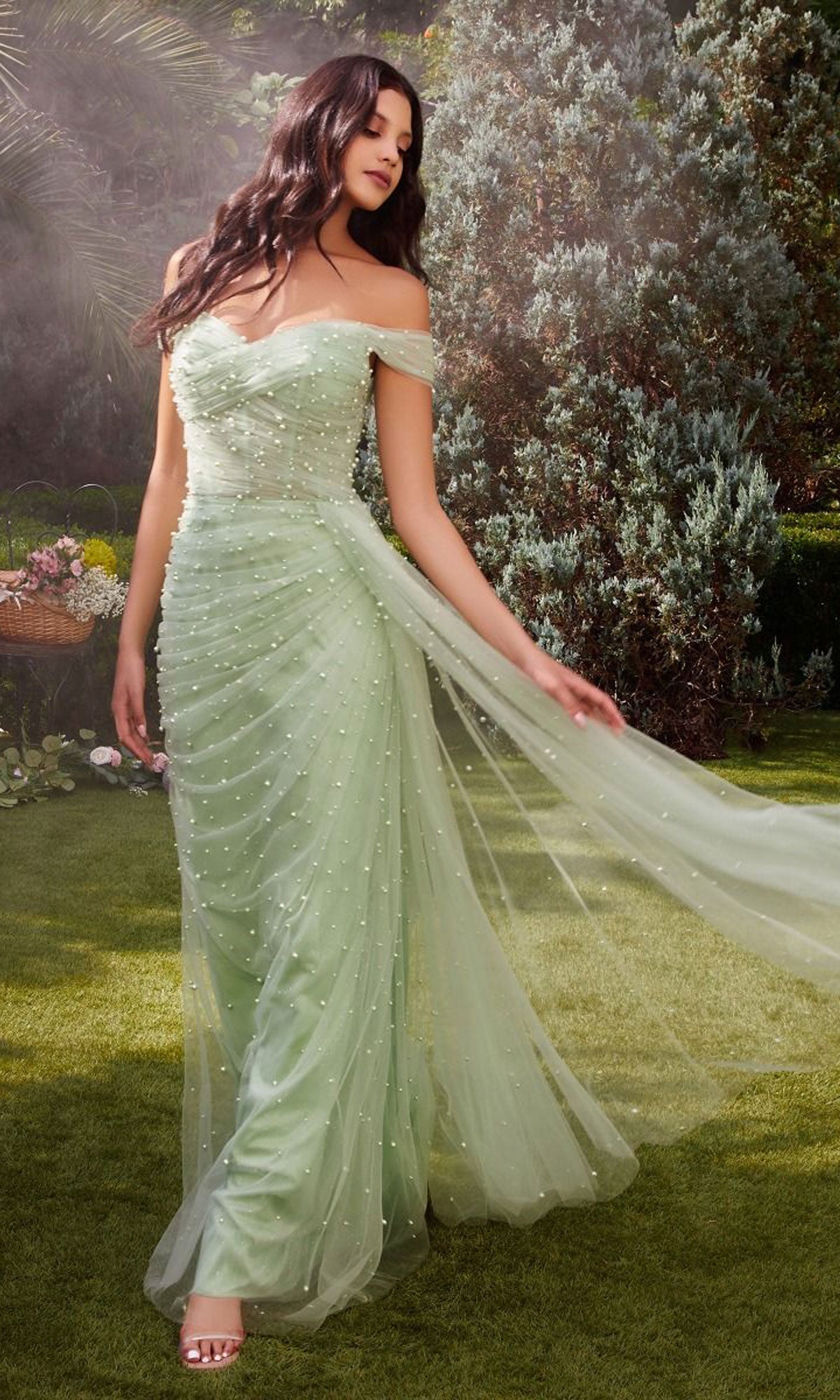 A woman in a long pink pearlized formal dress with a cape stands on grass, holding one side of the sheer cape, with greenery and flowers in the background.